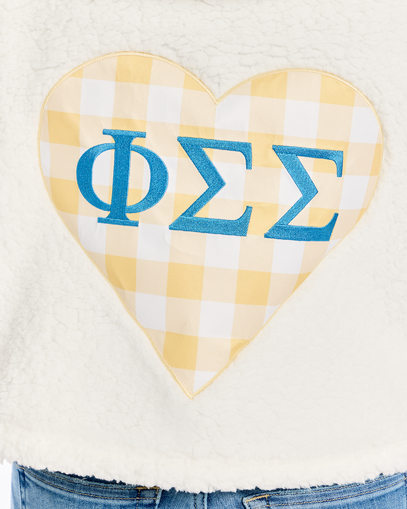 A close-up of a cream-colored fuzzy fleece garment featuring a large heart-shaped patch with a yellow and white gingham pattern and blue Greek letters 'ΦΣΣ' on it.