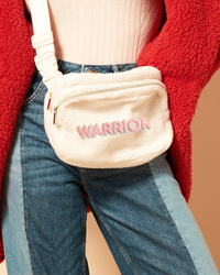 Beige fanny pack with 'Warrior' text worn by a person in a red jacket and blue jeans.