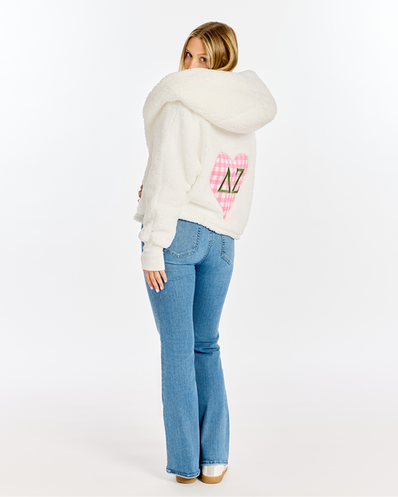 A woman shown from the back wearing a fluffy white hooded sweater with a pink plaid heart and Greek letters on the back, paired with blue jeans, against a plain white studio background.