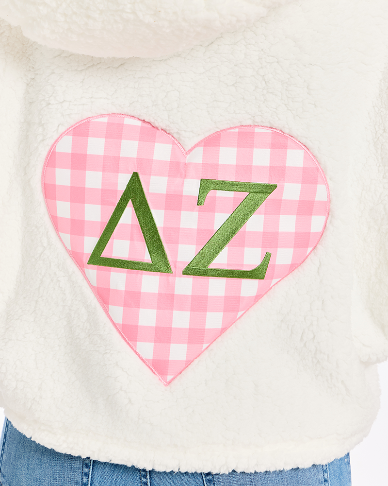 Close-up of a white sherpa hoodie featuring a pink and white gingham heart patch on the front with green Greek letters Delta Zeta.