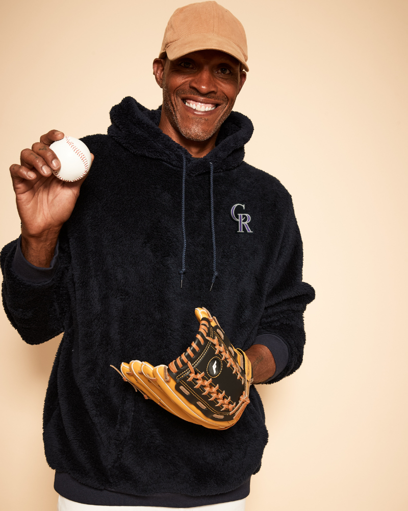 Man wearing a navy hoodie with a colorado rockies logo, holding a baseball and glove against a beige background