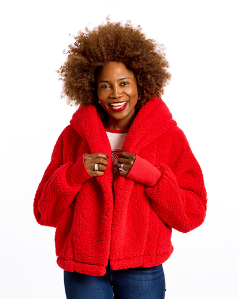A smiling woman with an afro hairstyle wearing a bright red sherpa fleece jacket, blue jeans, and a white top, photographed against a solid white background.