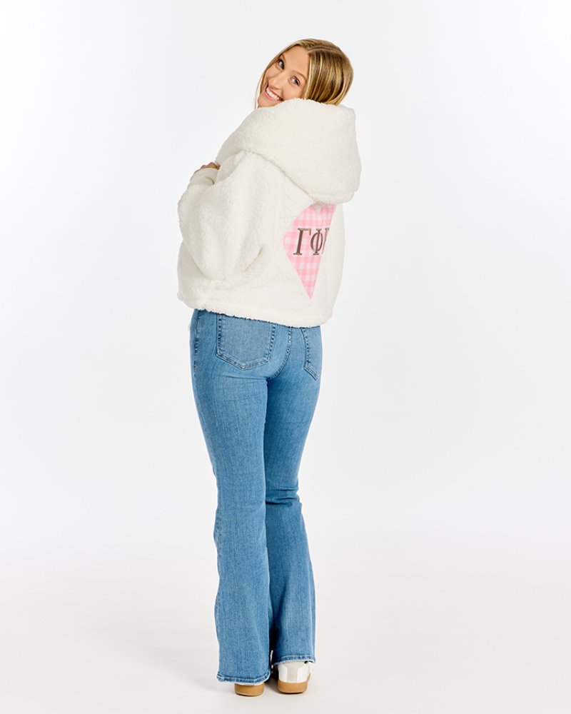 A woman wearing a white sherpa fleece cropped hoodie with a pink gingham heart and 'Gamma Phi Beta' greek letters on the back, paired with blue jeans, photographed against a plain white studio background.