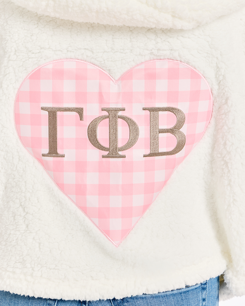 Close-up of a white sherpa fleece garment featuring a large pink gingham heart patch with metallic taupe Greek letters 'Gamma Phi Beta' on it.