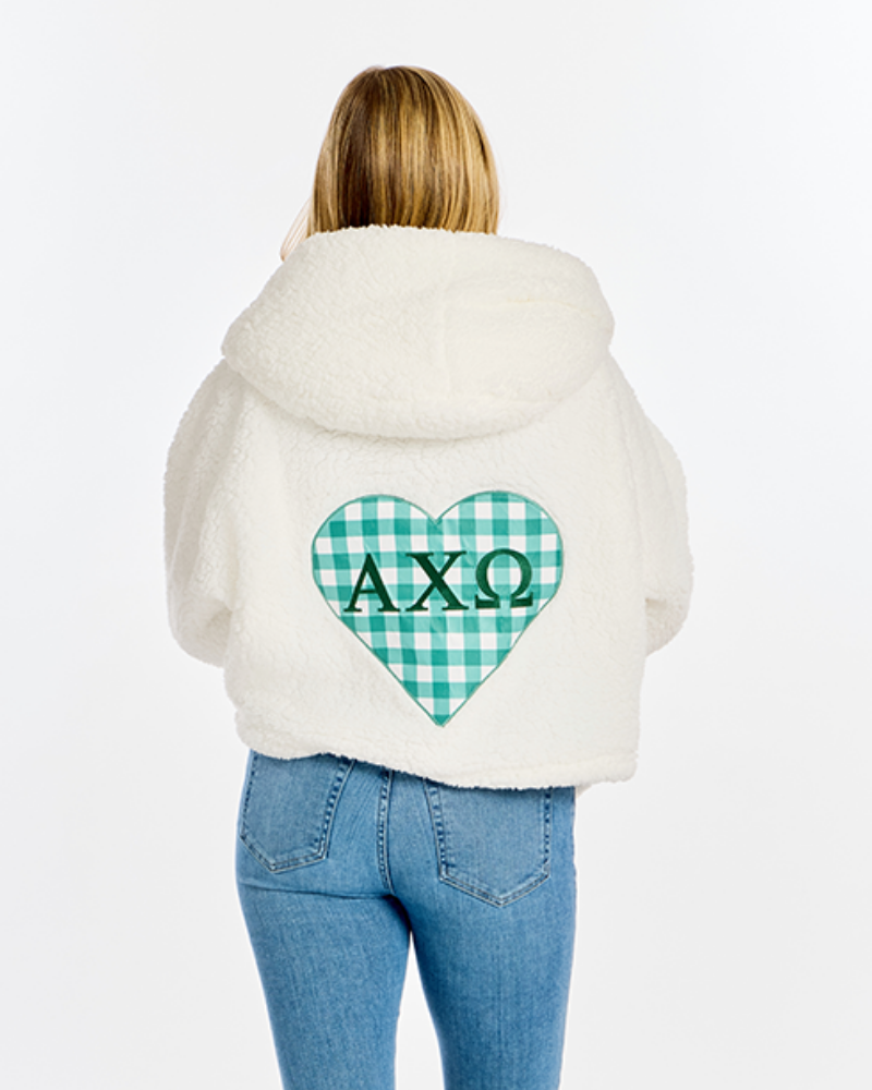 A woman viewed from behind wearing a cream sherpa hoodie with a green and white gingham heart graphic on the back featuring the Greek letters 'AXΩ'. She is also wearing blue jeans against a plain white background.