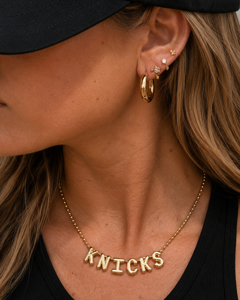 Close-up of a person wearing gold hoop earrings and a 'KNICKS' necklace.