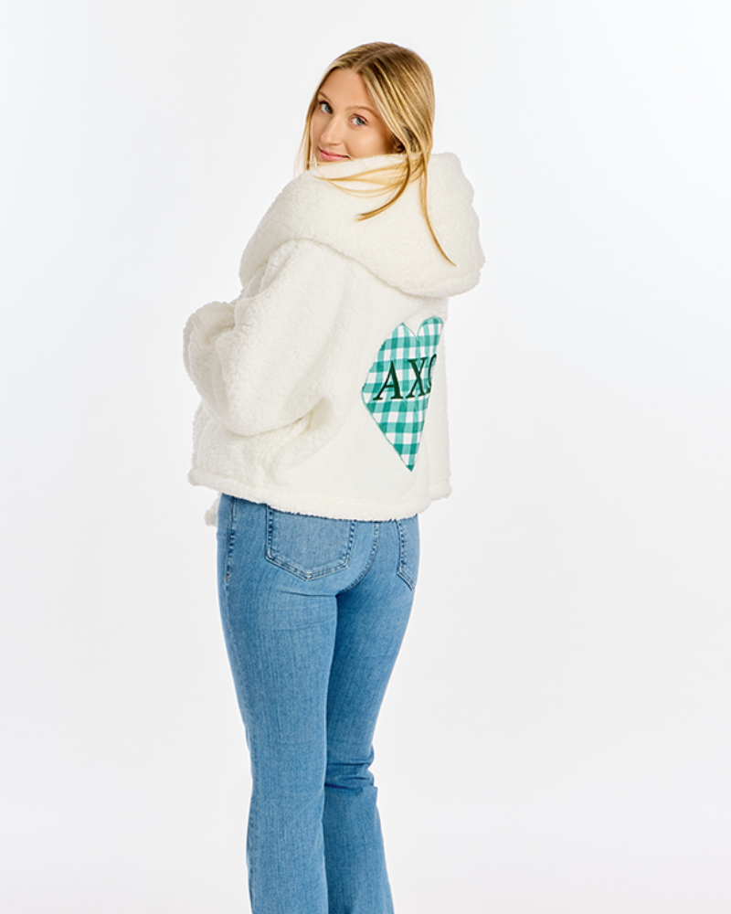 A woman wearing a white sherpa fleece hooded sweatshirt with a green and white gingham heart patch on the back, paired with blue jeans, photographed against a solid white background.