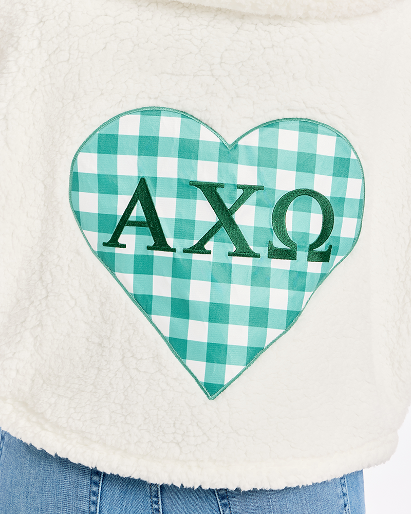 Close-up of a cream sherpa fleece garment with a large green-and-white gingham heart patch on the front featuring the Greek letters Alpha Chi Omega.
