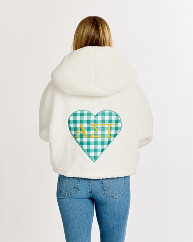 A person viewed from the back wearing a cream sherpa hoodie with a green and white gingham heart graphic on the back featuring the Greek letters Alpha Sigma Tau in gold, paired with blue jeans against a plain white background.