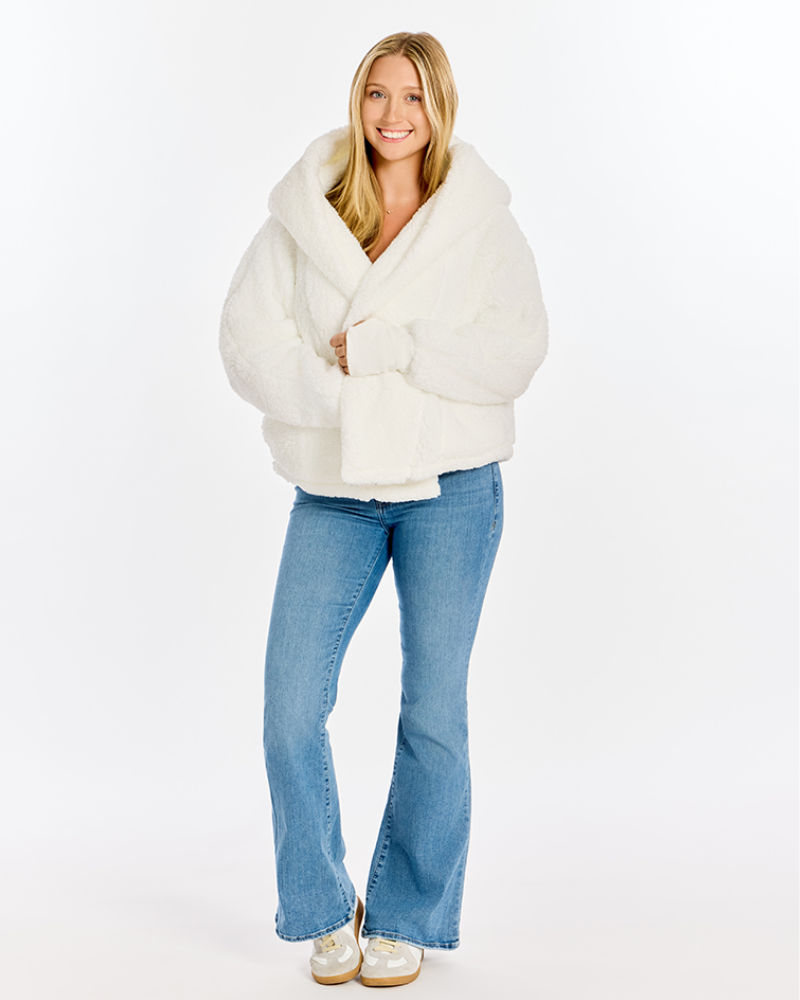 A woman modeling a fluffy white cropped sherpa jacket with a wide collar, paired with light blue flared jeans and white sneakers, against a plain white studio background.