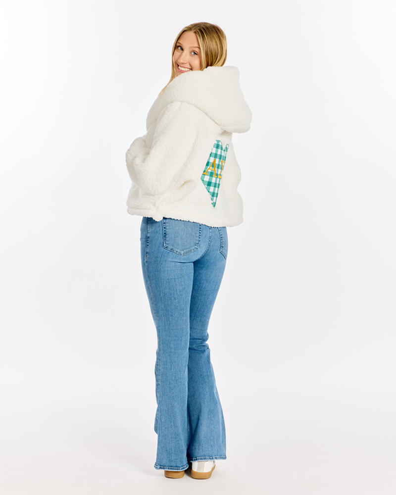 A woman wearing a white sherpa fleece hooded jacket with a green and yellow plaid accent patch on the back, paired with blue flared jeans, photographed from behind against a white studio background.