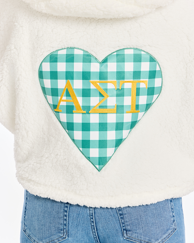 A close-up of a person wearing a cream sherpa hoodie with a large green and white gingham heart patch on the back featuring the Greek letters Alpha Sigma Tau in gold.