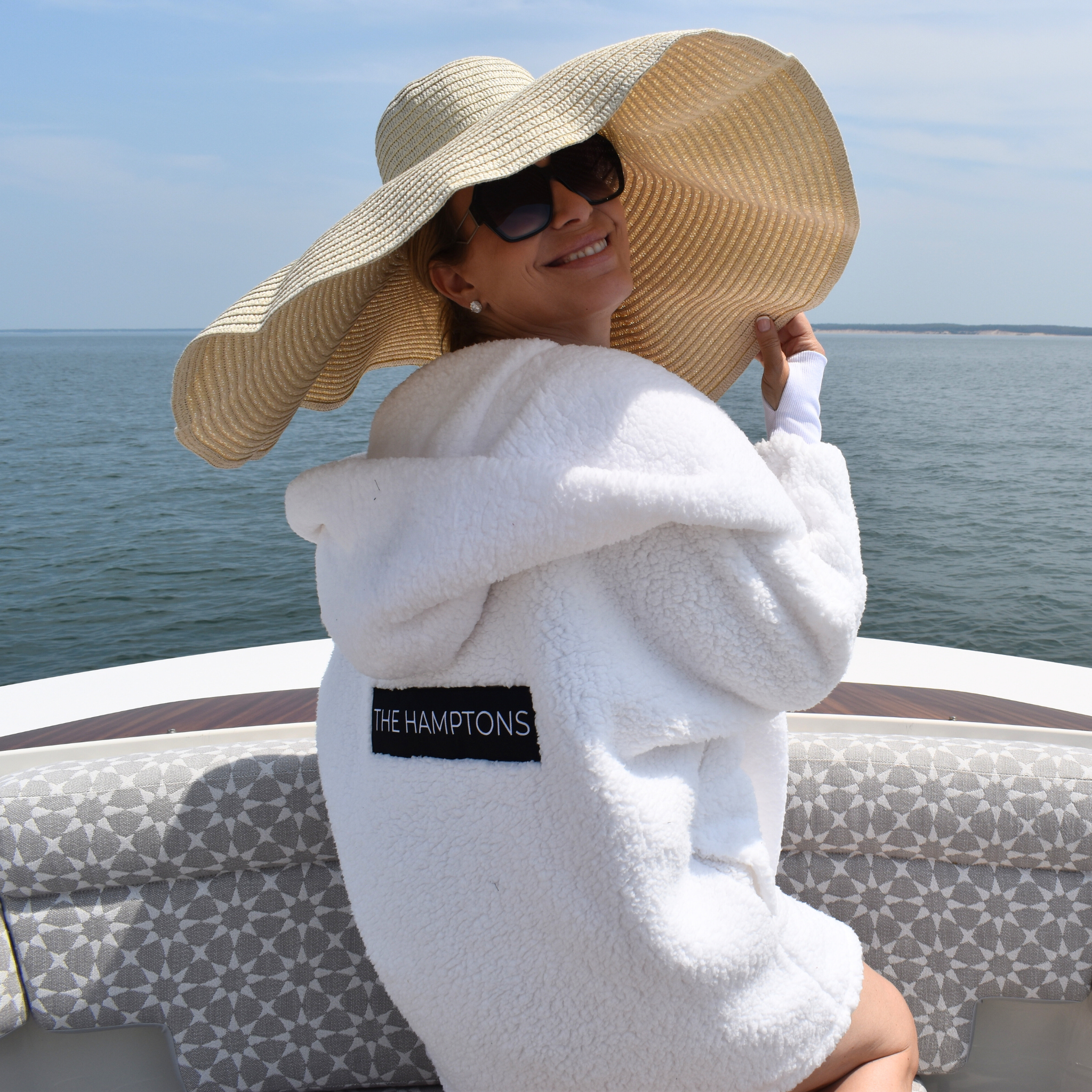 Person wearing a white lounger with 'The Hamptons' logo and a large sun hat, sitting on a boat.