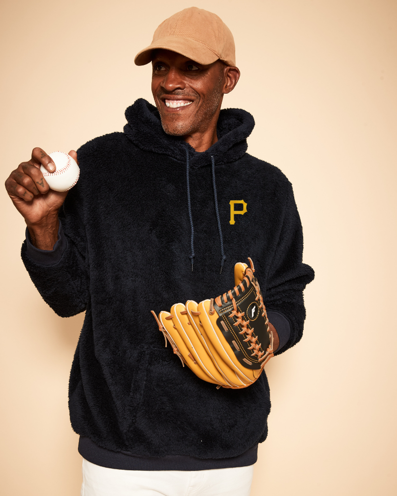 Man wearing a navy hoodie with a yellow pittsburgh pirates logo, holding a baseball and glove against a beige background
