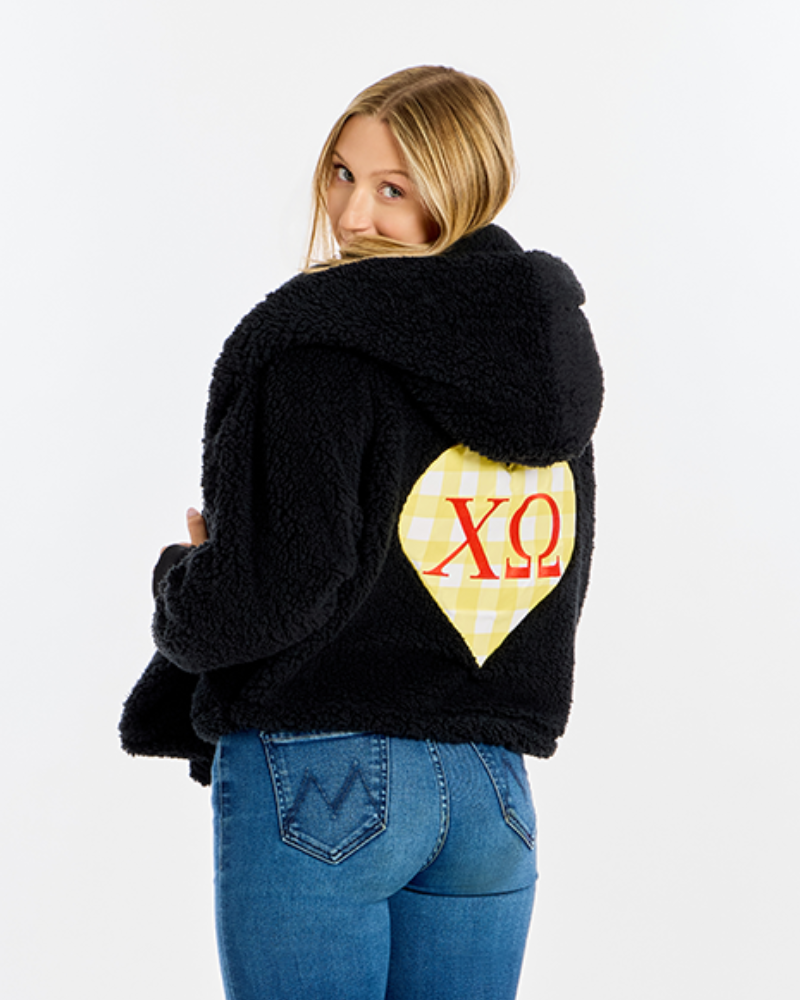 A woman wearing a black sherpa fleece hooded jacket with a yellow and white gingham heart patch on the back featuring red Chi Omega Greek letters, paired with blue jeans, photographed against a plain white background.