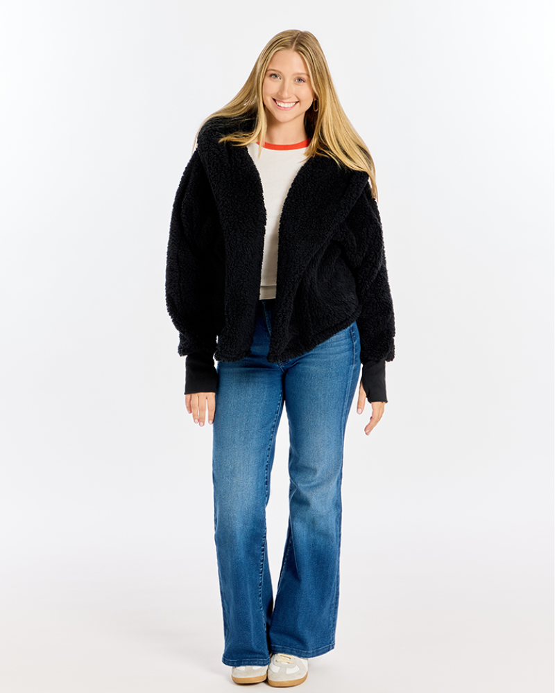 A woman modeling a black fuzzy cropped jacket over a cream top and blue jeans, standing against a plain white studio background.