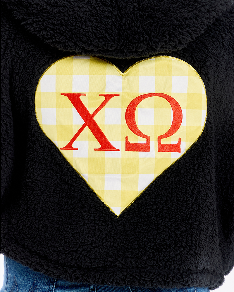 Close-up of a black sherpa hoodie featuring a large yellow and white gingham heart on the front with red Greek letters 'Chi Omega' in the center.