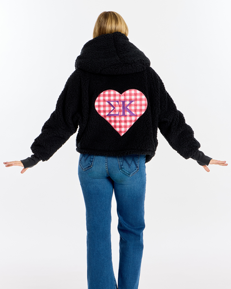 A model viewed from the back wearing a black sherpa hoodie with a large red and white gingham heart on the back featuring purple Greek Sigma Kappa letters, paired with blue jeans against a white studio background.