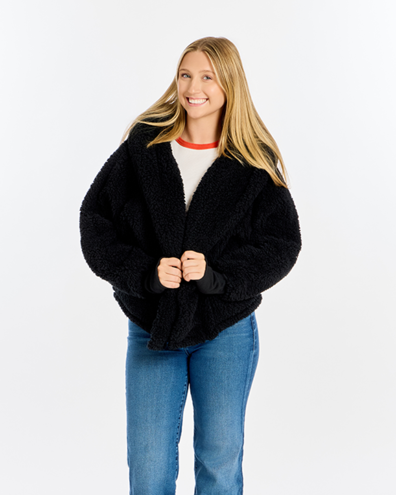 A woman modeling a black teddy fleece cardigan with a shawl collar, worn over a white crewneck top with a red collar trim and blue jeans, against a plain white studio background.
