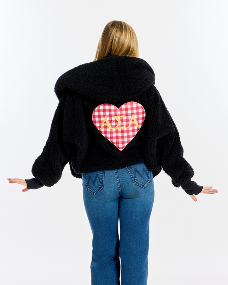 A woman viewed from behind wearing a black fuzzy hooded jacket with a red and white gingham heart patch on the back featuring the letters 'Alpha Sigma Alpha' in gold, paired with blue jeans against a white studio background.