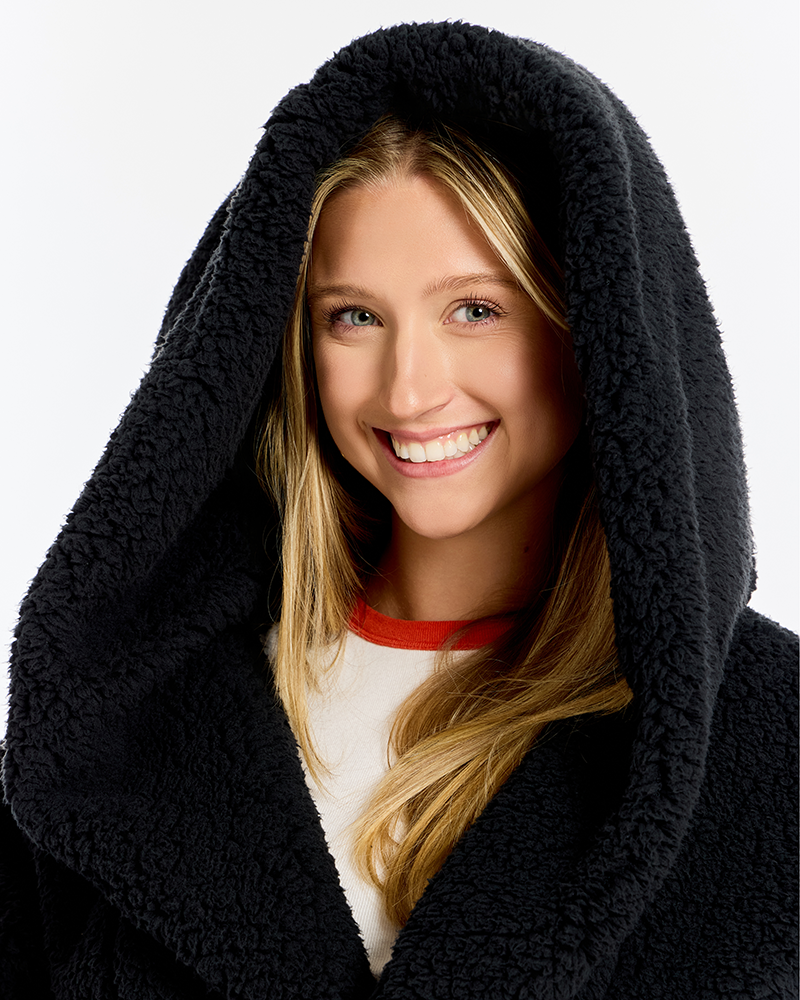A smiling blonde woman wearing a black sherpa fleece hooded robe or blanket, with the hood up, against a plain light gray studio background.