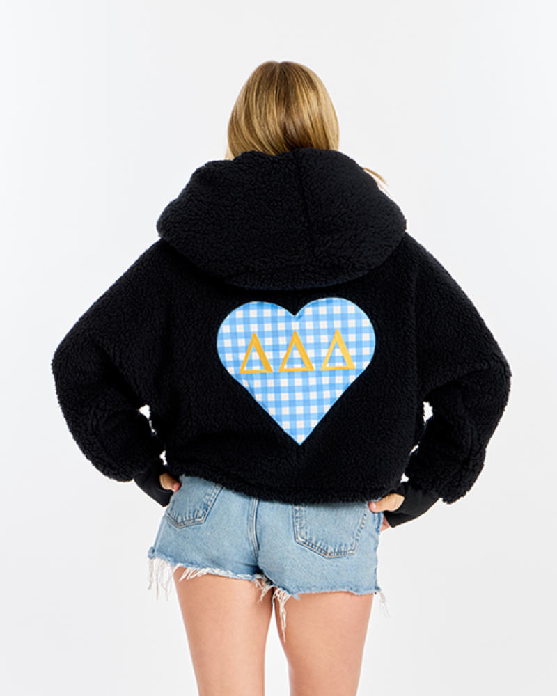 A woman viewed from behind wearing a black sherpa hoodie with a large blue-and-white gingham heart graphic on the back featuring three gold Greek letters, paired with light-wash denim shorts, against a plain white studio background.