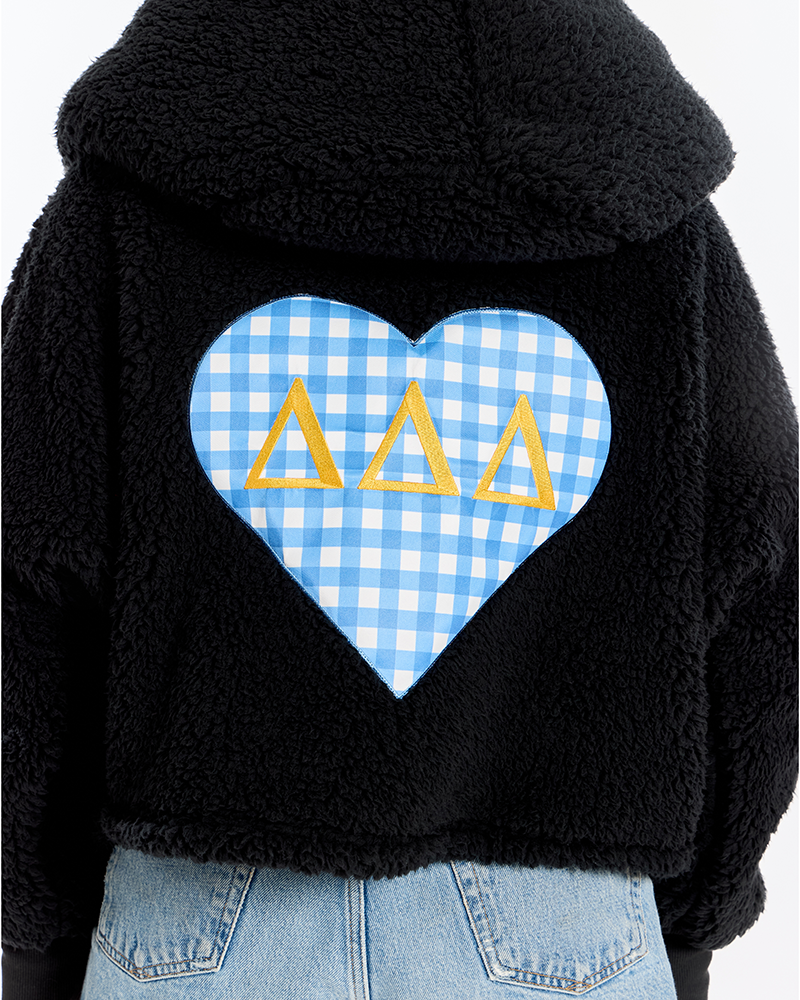 A close-up of a black sherpa fleece cropped hoodie from the back, featuring a large light blue and white gingham heart patch with three yellow Greek letters (Delta Delta Delta) in the center, worn with light wash blue jeans.
