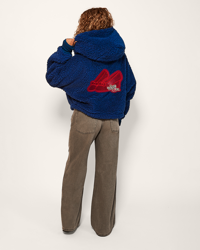 Person wearing a blue fleece jacket with a red logo on the back, standing against a white background