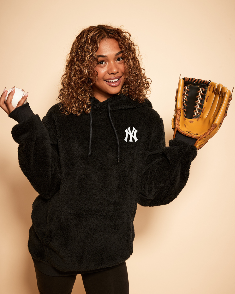 Person wearing a black hoodie with a new york yankees logo, holding a baseball and a glove against a beige background