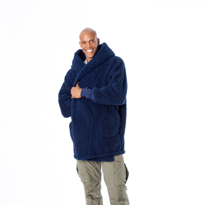 A person smiles warmly, wearing the Jill Martin University of Michigan Lounger—a large blue fuzzy hooded coat—paired with olive cargo pants against a plain white background, their arms crossed inside the oversized coat.