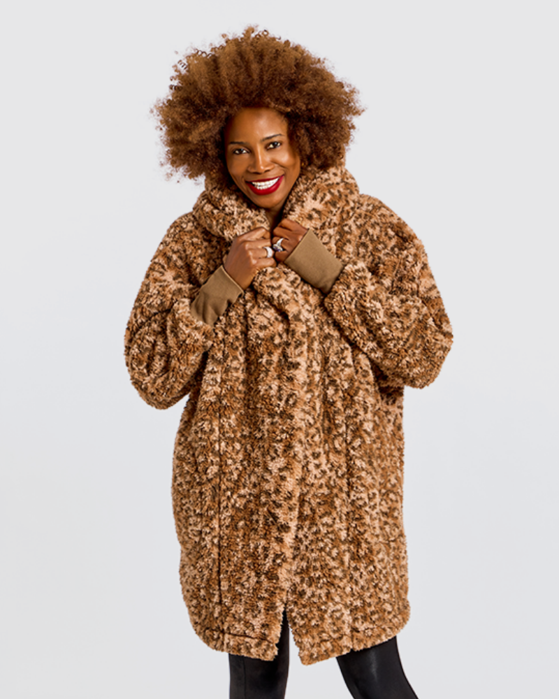 A smiling woman with an afro hairstyle wearing a brown and beige leopard-print faux fur coat with solid brown cuffs, photographed against a light gray studio background.|Model is 5'7" wearing size XS/S