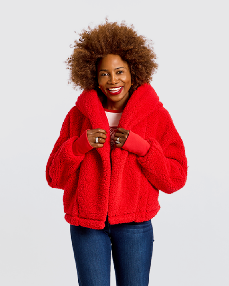A smiling woman with an afro hairstyle wearing a bright red sherpa fleece jacket, blue jeans, and a white top, standing against a light gray studio background.|Model is 5'7" wearing size XS/S