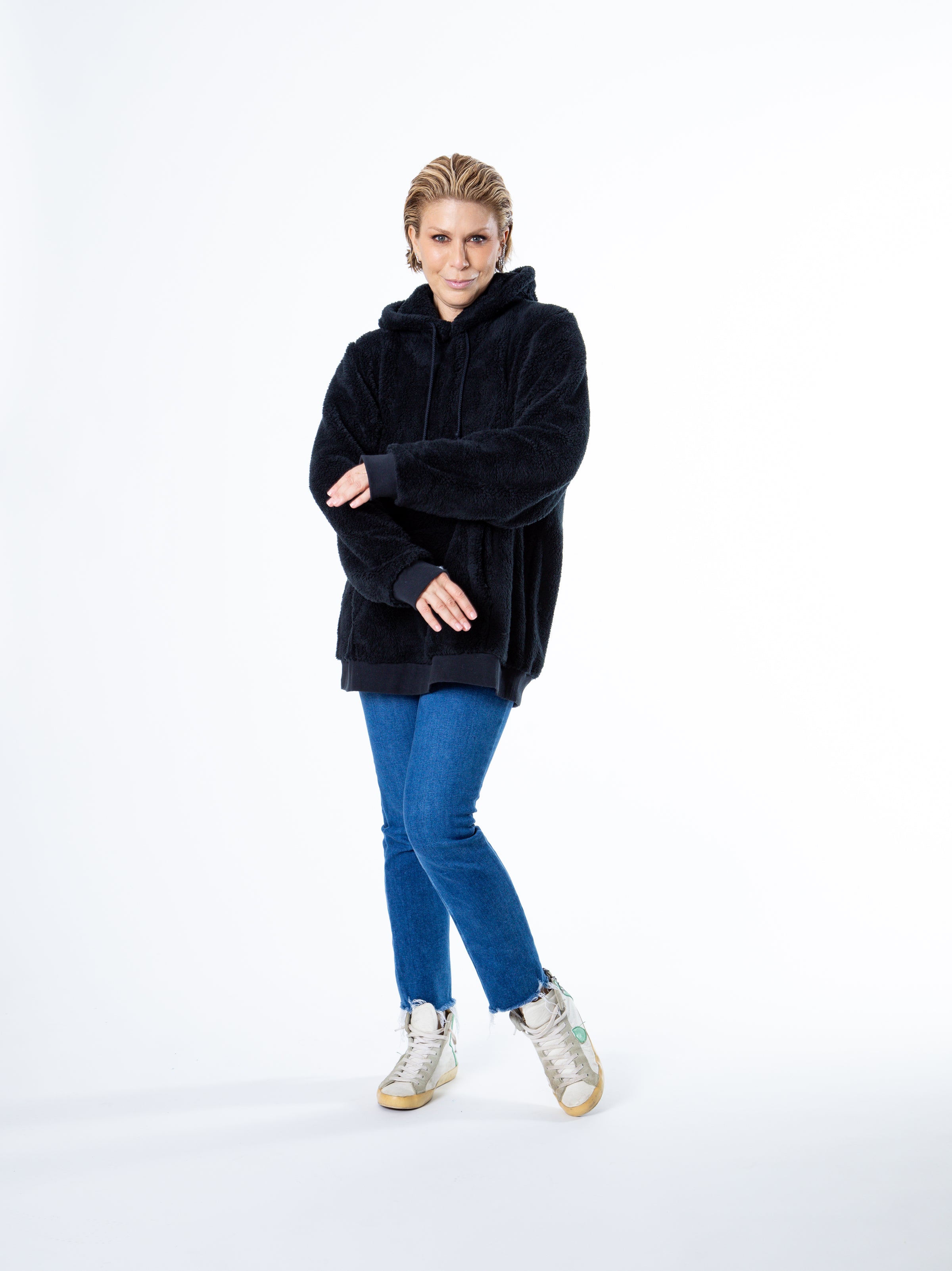Blonde woman wearing a black sherpa sweatshirt and skinny jeans, arms positioned in front of her body.