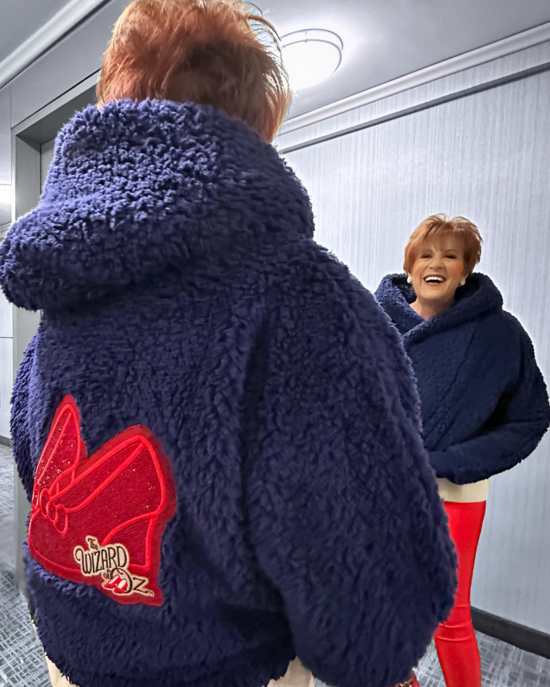 Person wearing a blue sherpa jacket with a ruby red slippers patch, standing in front of a mirror.