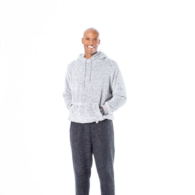 Against a plain white background, a person smiles and stands relaxed with hands in the pockets of their light gray Classic Hoodie by Jill Martin, dark gray sweatpants, and black sneakers.