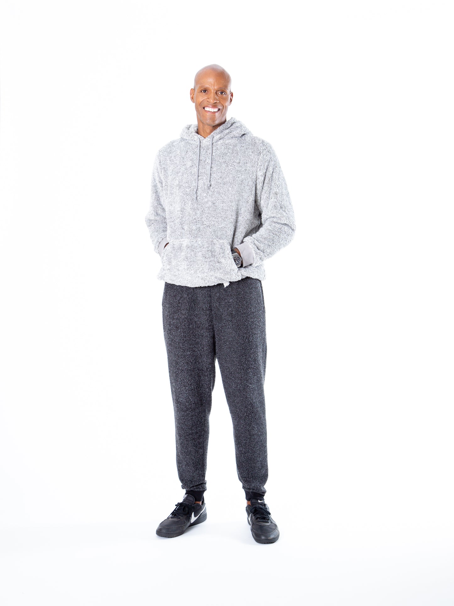 Against a plain white background, a person smiles and stands relaxed with hands in the pockets of their light gray Classic Hoodie by Jill Martin, dark gray sweatpants, and black sneakers.