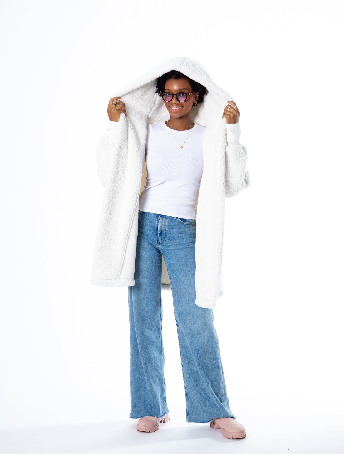 A person wearing glasses, a Jill Martin Classic Lounger in cream with the hood up, a white shirt, and blue jeans. They are smiling and posing against a plain white background.