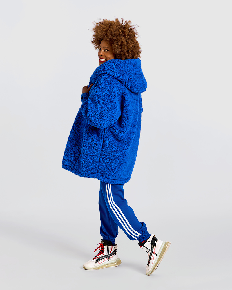 A child model wearing a bright blue sherpa fleece hooded jacket, matching blue track pants with white stripes, and white sneakers with red and black details, photographed against a light gray studio background.|Model is 5'7" wearing size XS/S