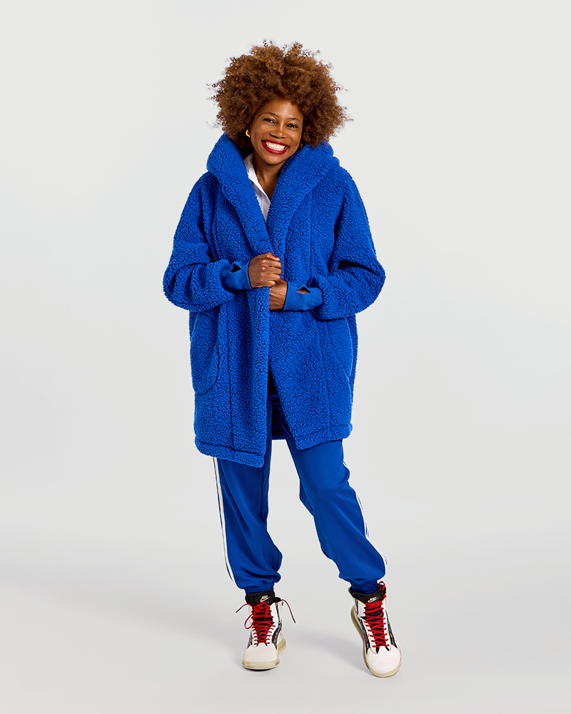 A woman modeling a vibrant royal blue sherpa fleece robe jacket, paired with matching blue track pants and white sneakers with red laces, against a solid light gray background.|Model is 5'7" wearing size XS/S