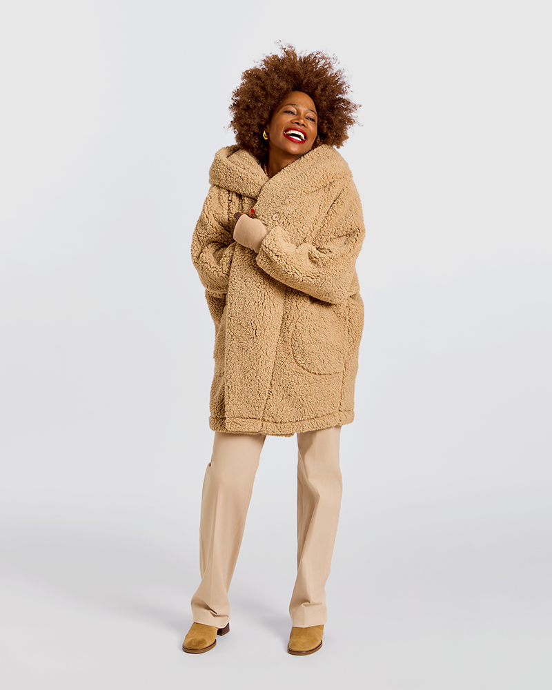 A woman modeling a cozy, oversized camel-colored teddy fleece coat with a wide collar and front patch pockets, paired with beige trousers and tan shoes, against a solid light gray background.|Model is 5'7" wearing size XS/S
