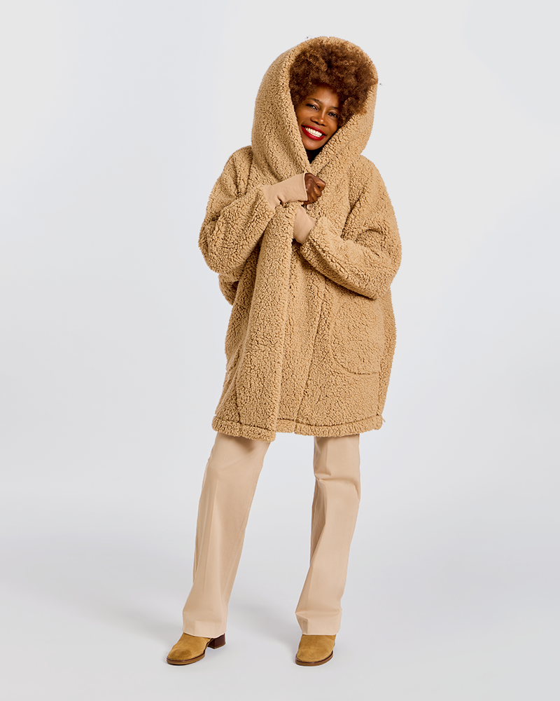 A woman modeling a cozy, oversized teddy fleece hooded coat in a warm camel tan color, paired with beige trousers and tan shoes, against a plain light gray studio background.|Model is 5'7" wearing size XS/S