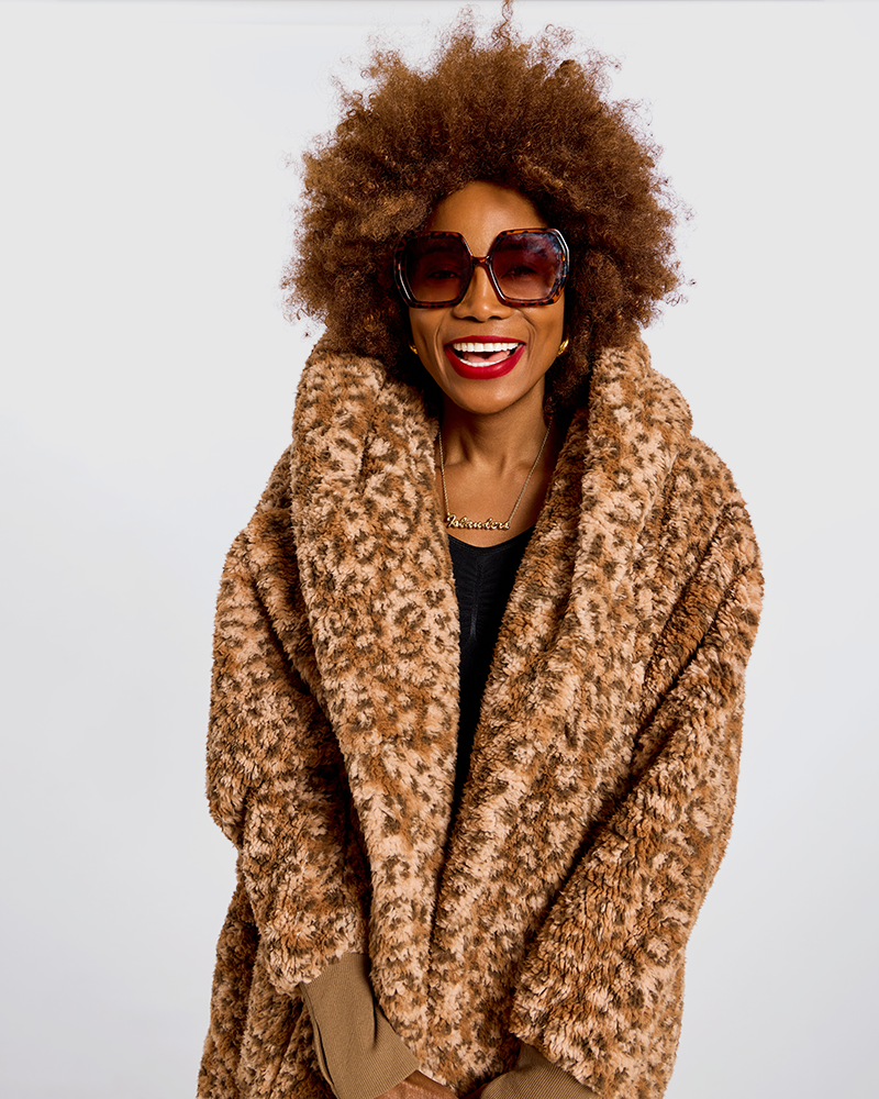 A woman with an afro hairstyle wearing oversized dark sunglasses and a brown and beige leopard-print faux fur wrap coat over a black top, photographed against a plain light gray background.|Model is 5'7" wearing size XS/S