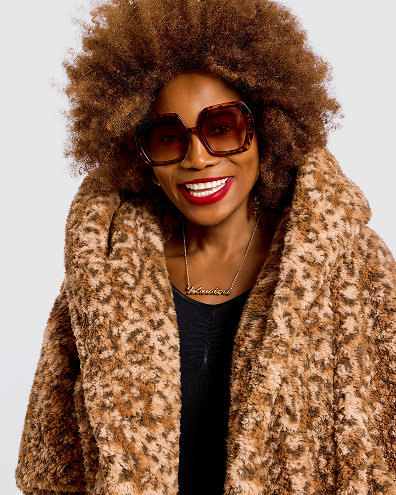 A smiling woman with an afro hairstyle wearing oversized tortoiseshell sunglasses and a plush leopard-print faux fur coat over a black top, photographed against a light gray studio background.|Model is 5'7" wearing size XS/S