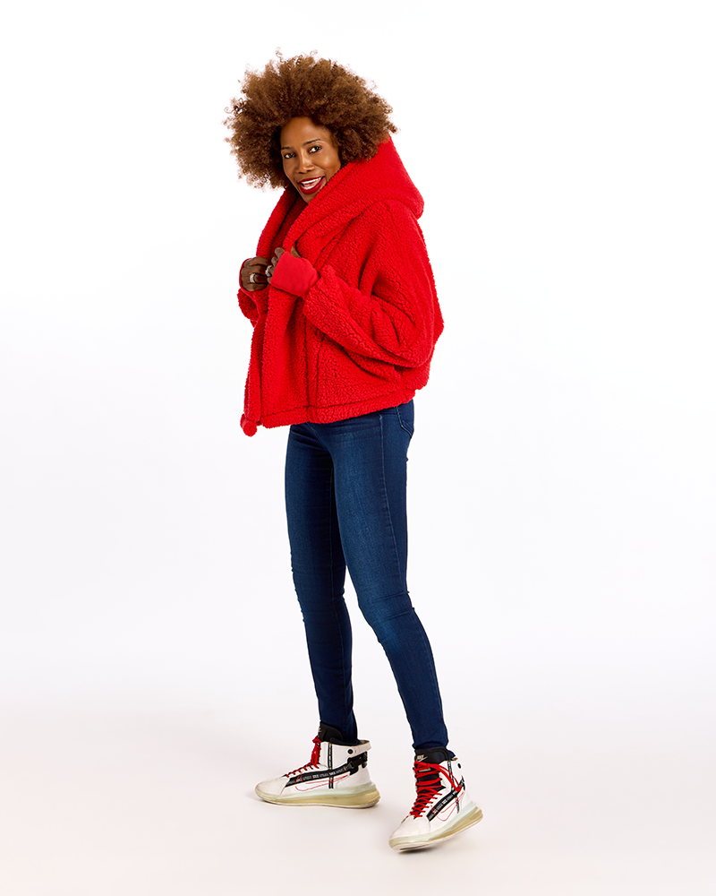 A woman modeling a bright red textured hooded jacket, paired with dark blue skinny jeans and white high-top sneakers with red and black details, against a solid white studio background.