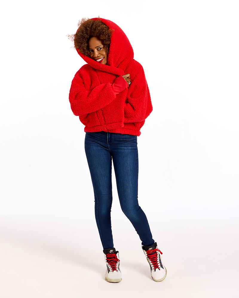 A woman modeling a bright red fuzzy hooded sweatshirt, paired with dark blue skinny jeans and red-and-white sneakers, against a plain white studio background.