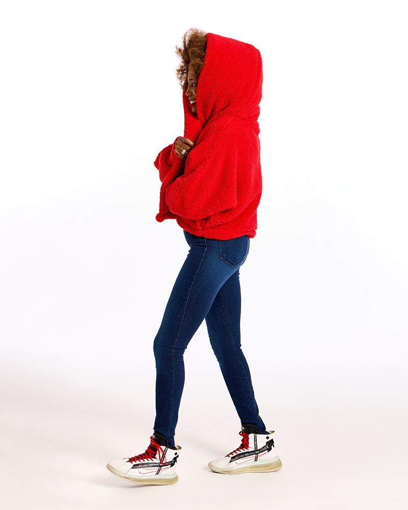 A person shown in side profile wearing a bright red hooded sweatshirt with brown faux-fur trim, dark blue skinny jeans, and white high-top sneakers with red and black details, photographed against a clean white studio background.