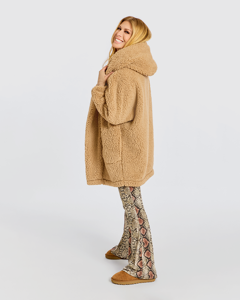 A woman modeling a camel-colored sherpa fleece hooded coat, paired with snakeskin-patterned flared pants and tan slippers, against a plain light gray studio background.|Jill is 5'6" wearing size XS/S