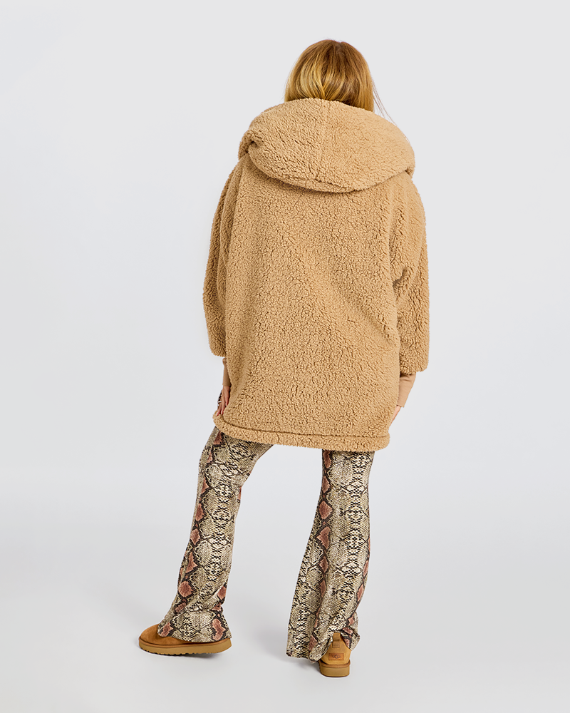 A model shown from the back wearing a camel-colored sherpa fleece hooded jacket, patterned snakeskin-print pants, and tan boots, set against a plain light gray studio background.|Jill is 5'6" wearing size XS/S