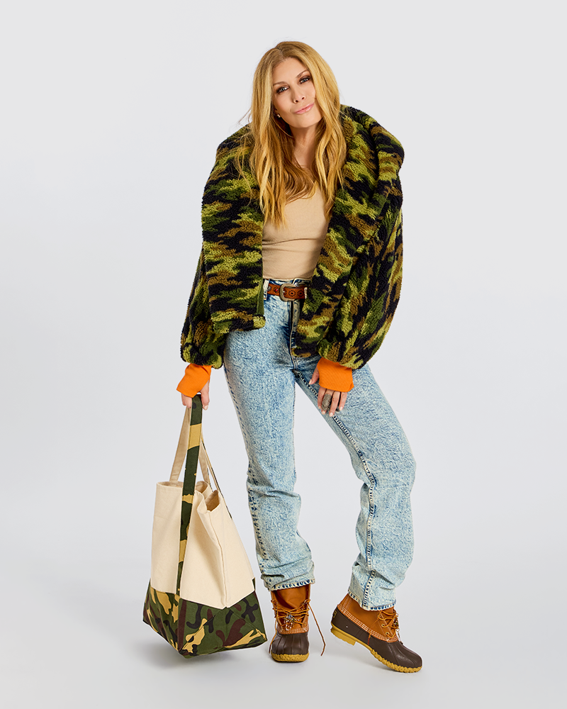 A model stands against a light gray studio background wearing a fuzzy green and black camouflage jacket, a beige top, light blue jeans, and brown lace-up boots, while holding a matching camouflage tote bag.|Jill is 5'6" wearing size XS/S