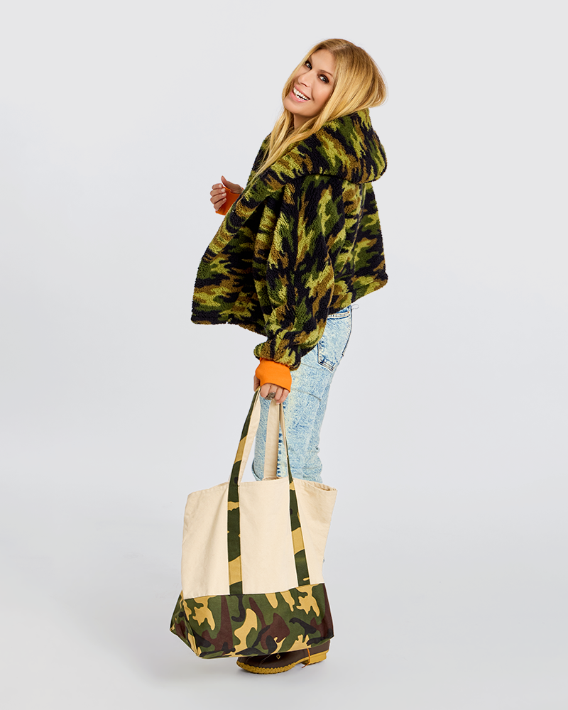 A woman wearing a green and black camouflage hooded jacket, light blue jeans, and orange cuffs is holding a matching camouflage and canvas tote bag against a light gray studio background.|Jill is 5'6" wearing size XS/S